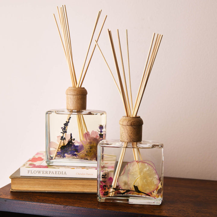 Rosy Rings Apricot Rose Scented Reed Diffuser with 10 reed sticks and wooden cap displayed on top of wooden shelf and flower books