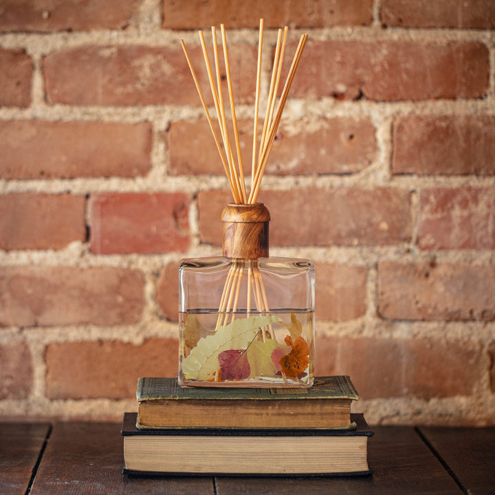 Rosy Rings Honey Tobacco Scented Reed Diffuser with 10 reed sticks and wooden cap displayed on top of wooden shelf and flower books