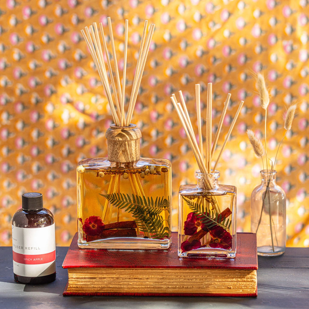 Rosy Rings Spicy Apple Scented Reed Diffuser with 10 reed sticks and wooden cap displayed on top of wooden shelf and flower books